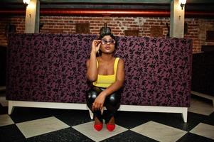 Stunning African American women in yellow top and black leather pants, sunglasses, pose at pub. photo
