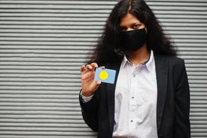 mujer asiática con ropa formal y máscara protectora negra sostiene la bandera de palau a mano contra el fondo gris. coronavirus en el concepto de país. foto