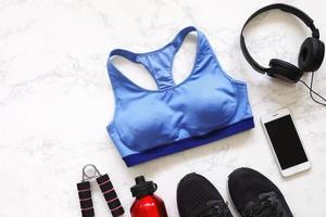 Top view of flat lay fitness equipment with sport bra, sneakers shoes, smartphone, earphones and bottle of water on white marble background photo