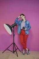 Young woman leaning against studio flashlight and holding camera over a pink background photo