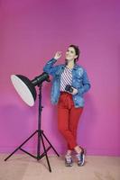 Young woman leaning against studio flashlight and holding camera over a pink background photo