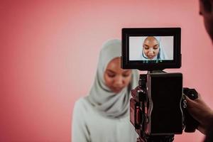 Videographer in digital studio recording video on professional camera by shooting female Muslim woman wearing hijab scarf plastic pink background. photo