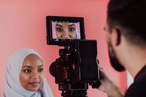 Videographer in digital studio recording video on professional camera by shooting female Muslim woman wearing hijab scarf plastic pink background. photo