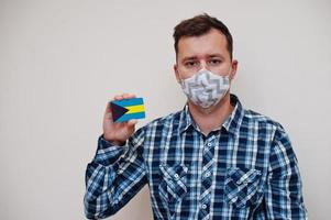 Man in checkered shirt show The Bahamas flag card in hand, wear protect mask isolated on white background. American countries Coronavirus concept. photo