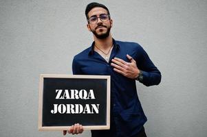 el hombre árabe usa camisa azul y anteojos sostiene una tabla con la inscripción zarqa jordan. ciudades más grandes en el concepto del mundo islámico. foto