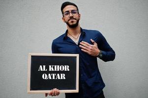 Arab man wear blue shirt and eyeglasses hold board with Al Khor Qatar inscription. Largest cities in islamic world concept. photo