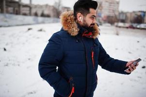 Beard indian man wear jacket in cold winter day. He speak mobile phone by wireless headphones. photo