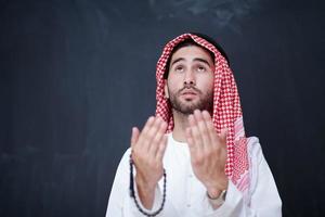 arabian man making traditional prayer to God, keeps hands in praying gesture in front of black chalkboard photo
