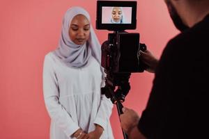 videógrafo en estudio digital grabando video en una cámara profesional disparando a una mujer musulmana con bufanda hiyab de fondo rosa de plástico. foto