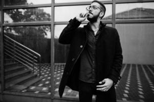 Middle eastern entrepreneur wear black coat and blue shirt, eyeglasses against office building smoking cigarette. photo