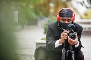 joven camarógrafo afroamericano profesional con cámara profesional con equipo profesional. camarógrafo afro con duraq negro y máscara de protección facial, haciendo videos. foto