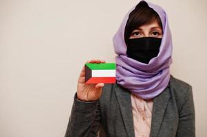 Portrait of young muslim woman wearing formal wear, protect face mask and hijab head scarf, hold Kuwait flag card against isolated background. Coronavirus country concept. photo