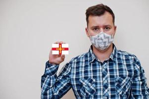 Man in checkered shirt show Bailiwick of Guernsey flag card in hand, wear protect mask isolated on white background. Europe countries Coronavirus concept. photo