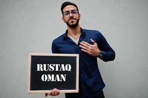 Arab man wear blue shirt and eyeglasses hold board with Rustaq Oman inscription. Largest cities in islamic world concept. photo