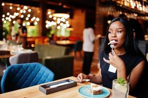 Fashionable feminist african american woman wear in black t-shirt and shorts, posed at restaurant, eat cheese cake. photo