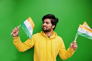 un joven indio hipster urbano con una sudadera amarilla de moda sostiene banderas de la india en las manos. Un chico fresco del sur de Asia usa una sudadera con capucha aislada en un fondo verde. foto