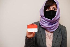 retrato de una joven musulmana que usa ropa formal, protege la máscara facial y el pañuelo de la cabeza del hiyab, sostiene la tarjeta de la bandera de indonesia contra un fondo aislado. concepto de país coronavirus. foto