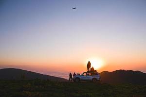 Group of travelers content creators stand by 4wd vehicle together outdoors in nature adventure watch sunset over horizon over cloudscape in wilderness. Exploration and adventure photo