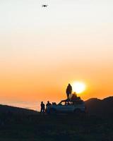 Group of travelers content creators stand by 4wd vehicle together outdoors in nature adventure watch sunset over horizon over cloudscape in wilderness. Exploration and adventure photo