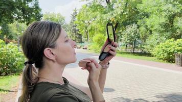 Middle-aged woman adjusts makeup on her face. She sits in park and holds in her hands small mirror with powder. video