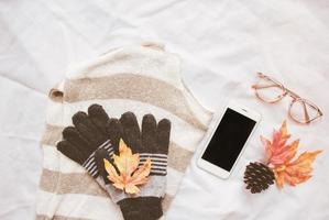 concepto de estilo de moda de otoño, suéter y teléfono inteligente con hojas de arce sobre fondo de sábana blanca foto