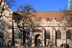 In December 2020, the cathedral in Augsburg stands behind bare trees and against a blue sky, with some people resting in the sun photo