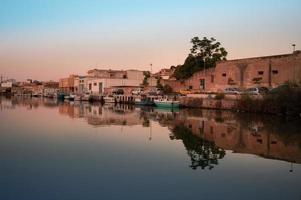 The sun is rising at the port of Mazara del Vallo in Sicily. The sky is reflected in the water. Houses line the harbor edge and boats are moored on the shore. photo