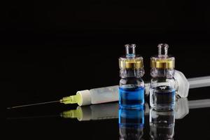 A syringe with a needle, an ampoule with a transparent liquid and an ampoule with a blue liquid are reflected against a dark background photo