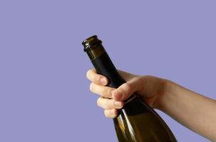 A woman's hand holding an open bottle of champagne on a lilac background.Very peri photo