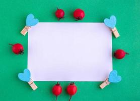 A white sheet of paper on a green background with red berries and clothespins with blue hearts. Holiday concept Valentine's Day, Mother's Day, Father's Day, New year. Space for your text. photo