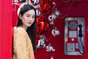 hermosa mujer asiática de pelo largo con una túnica amarilla y sonriendo alegremente parada frente a una cabina de teléfono roja con el tema de celebrar la navidad y feliz año nuevo foto