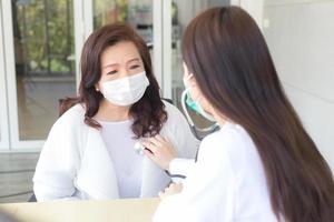 Asian woman doctor uses stethoscope to check health of lungs and heart or diagnosis symptom of woman patient while both wear face mask in hospital. photo