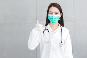 Asian woman doctor who wears medical coat and face mask shows thump up as good sign in health protection concept at hospital. photo