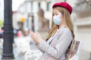 Asian beautiful girl wearing suit and red cap holds smartphone in her hands while sits on bench in park while wearing medical face mask in new normal lifestyle concept. photo