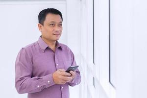 Asian business middle-age man is holding smartphone mobile phone in his hand and look out from window in the room. photo