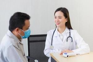 Asian beautiful young woman doctor talking with a man patient about his pain and symptom while they put on a face mask to prevent Coronavirus disease and Thermometer on table in hospital. photo