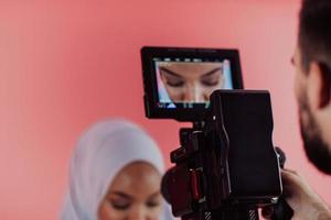 Videographer in digital studio recording video on professional camera by shooting female Muslim woman wearing hijab scarf plastic pink background. photo