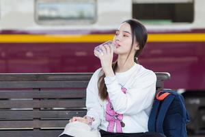 Asian beautiful woman who wears white long sleeve sits on chair and feels tried from traveling, so, she drinks water while waiting to go home at train station. photo