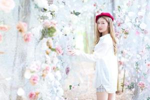 hermosa mujer asiática tiene el pelo largo de bronce y usa gorra roja, vestido blanco y botas negras mientras se para entre el jardín de flores en la temporada de invierno. foto