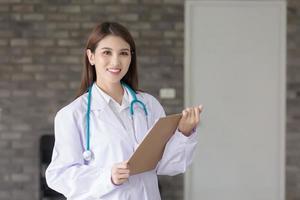 Asian woman doctor with a white lab coat stand and hold a clipboard in her hand at work office in hospital. Coronavirus protection concept. photo