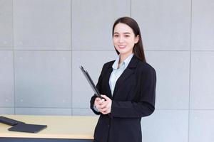 una joven trabajadora profesional asiática con traje negro sostiene un portapapeles en sus manos y sonríe confiada en la oficina. foto