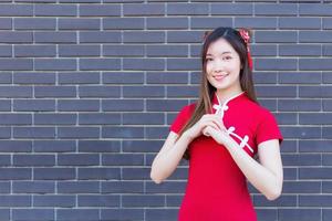 hermosa mujer asiática con cabello largo que usa un vestido rojo cheongsam en el tema del año nuevo chino mientras muestra su mano como saludo chino con una pared de ladrillo como fondo. foto