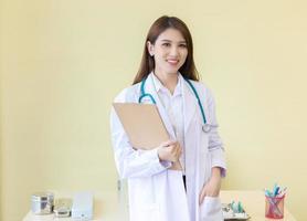 Young beautiful Asian woman doctor working at hospital. She wear a white robe and stethoscope and hold a clipboard in her hands. photo