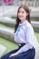 Beautiful Asian high school student girl in the school uniform with braces on her teeth sitting and smiles confidently while she looks at the camera happily with the building in the background. photo