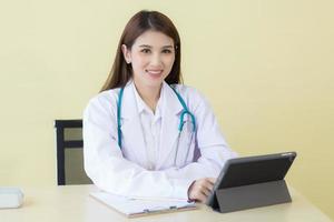 Professional Asian beautiful young smiling female doctor sitting in office at hospital. On table has a clipboard and digital tablet computer. photo