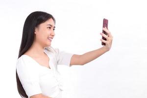 Portrait of Asian beautiful lady who has black long hair in white shirt, is holding the smartphone in her hand and smiling. She take a video conference on white background. photo