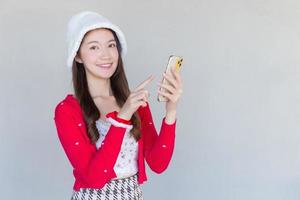 retrato de una linda adolescente asiática con un vestido rojo y un sombrero blanco que sonríe alegremente usando un smartphone con un fondo blanco. foto