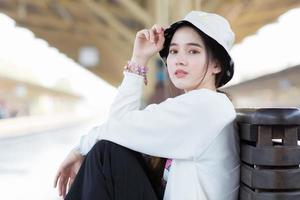 hermosa chica asiática con camisa blanca de manga larga y sombrero sentada en la estación de tren para esperar que llegue el tren. foto