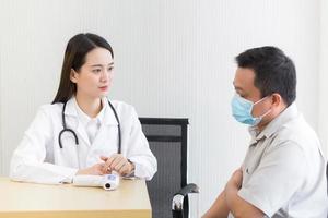 Asian beautiful woman doctor talking with a man patient about his pain and symptom while they put on a face mask to prevent Coronavirus disease and Thermometer on table in hospital. photo