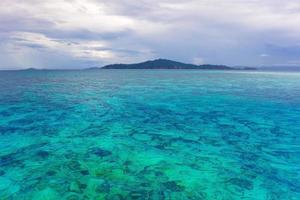 Mostly cloudy day with blue green ocean which we can see corals on sea ground and mountain as background. photo